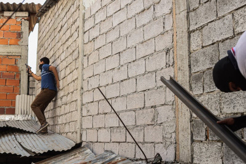A person in a blue shirt and face covering holds a gun while standing on a corrugated metal roof beside a brick wall. Another person crouches nearby, partially obscured by the wall and roof.