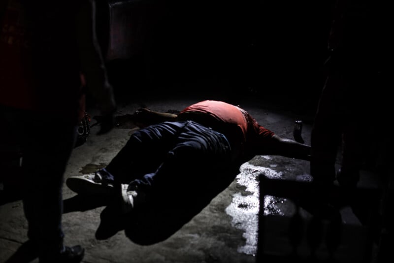 A person lies face-up on the ground in a dark, dimly lit area, with shadows obscuring surrounding details. The person appears motionless, wearing a red shirt and dark pants. Other figures are partially visible nearby.