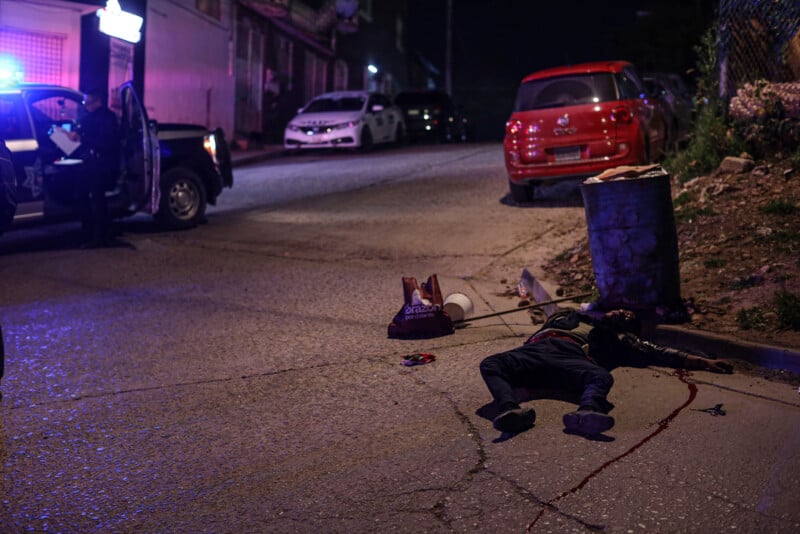 A person lies motionless on a street at night with a pool of blood near their head. Police cars with flashing lights and an officer are visible in the background. Groceries are scattered on the ground.