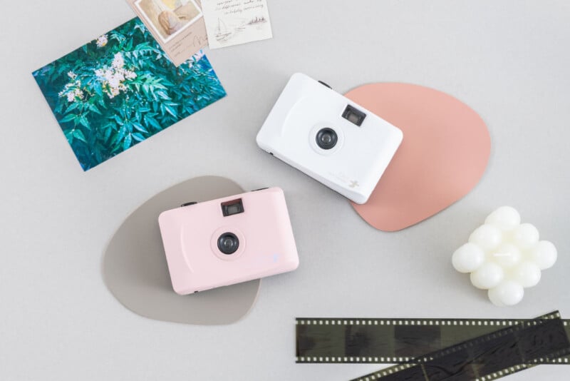 Dos cámaras de estilo retro, una rosa y otra blanca, colocadas sobre un bloque de color abstracto junto a postales, fotografías de hojas verdes, velas decorativas y una tira de película sobre una superficie gris claro.