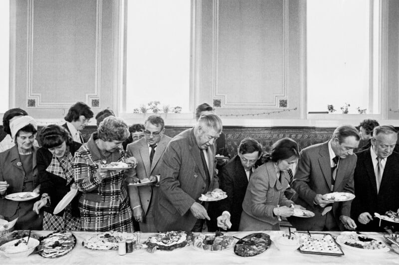 A group of people in formal attire serve themselves food from a buffet table at a social gathering in a large, elegant room with tall windows and ornate walls.