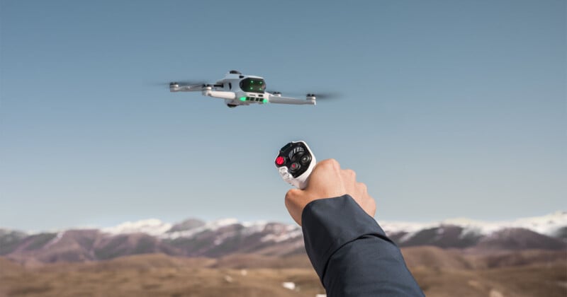 Un hombre que sostiene un control remoto opera un pequeño dron blanco que vuela sobre un paisaje marrón bajo un cielo despejado con montañas nevadas al fondo.