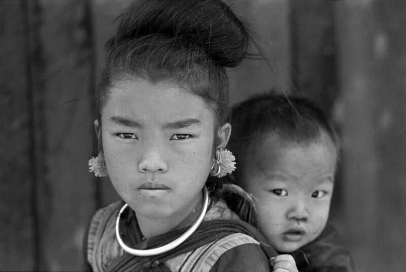 Una niña con flores en el cabello y un gran collar estaba parada al frente con una expresión seria en su rostro, mientras un niño pequeño se asomaba desde una bolsa de tela en su espalda. La imagen está en blanco y negro.