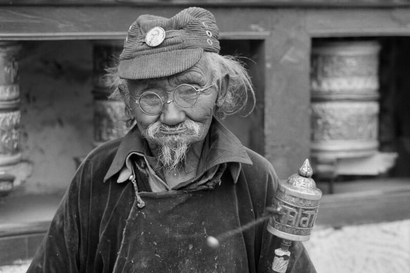 Un anciano con gafas, sombrero y bigote sostiene una rueda de oración frente a una gran rueda de oración giratoria. La imagen es en blanco y negro y los personajes van vestidos con ropa tradicional.