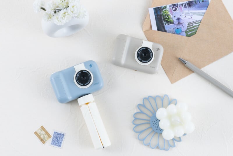 Dos cámaras de juguete, una azul y otra beige, están colocadas sobre una superficie blanca junto a una fotografía, un sobre, un bolígrafo, una vela floral blanca, un pequeño jarrón y dos sellos.