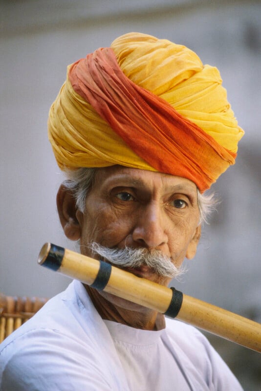 Un anciano de barba blanca lleva un turbante de color amarillo brillante y naranja. Con una camisa blanca, se llevaba una flauta de madera a los labios y parecía estar tocando. El fondo es suave y borroso.
