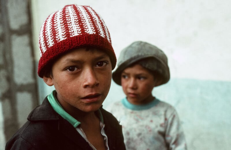 Había dos niños pequeños cerca de la pared. El niño en primer plano, con un gorro de punto rojo y blanco, mira a la cámara, mientras que el niño al fondo, con un gorro gris, mira hacia otro lado.