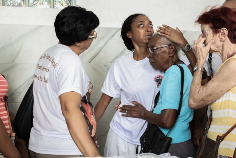 A grieving woman is comforted by others in a group, some embracing her and touching her head, inside a room with white marble walls. The mood is emotional and solemn.