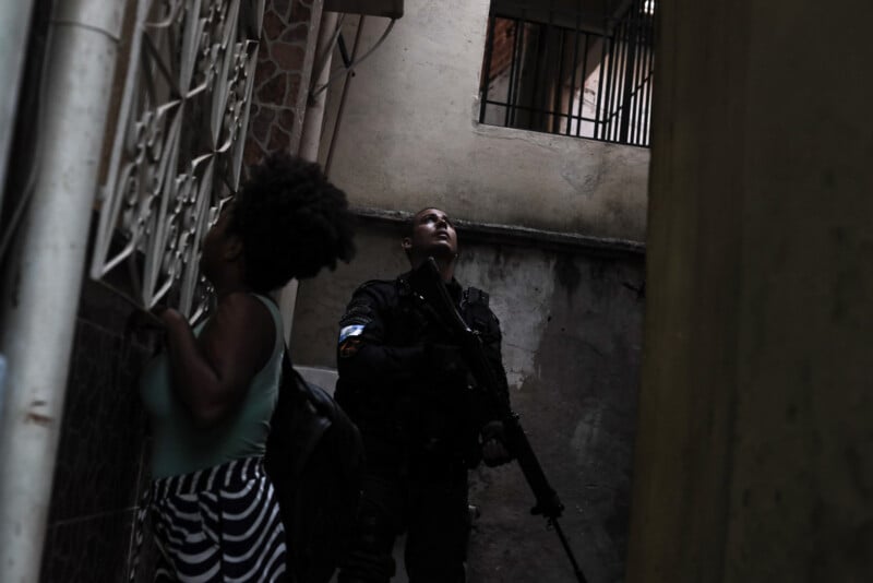 A police officer holding a rifle stands alert in a narrow alley, looking upward. In the foreground, a woman with curly hair enters a building, partially turned away from the camera. The setting appears tense and confined.