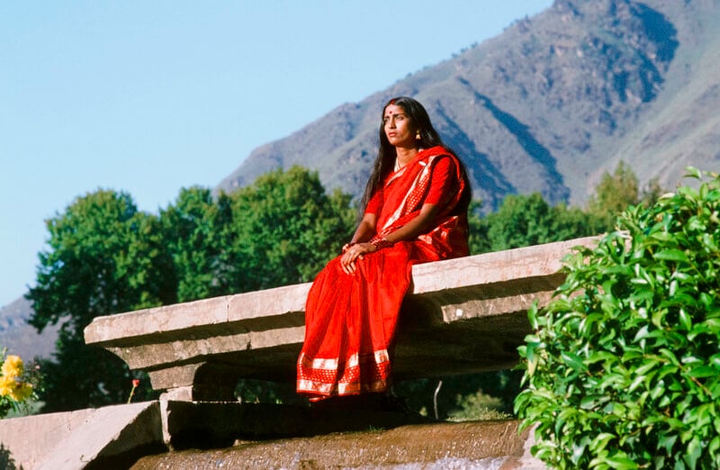 Una mujer vestida con un sari rojo brillante está sentada en un estante de piedra al aire libre, rodeada de vegetación, con colinas cubiertas de árboles y un cielo azul claro al fondo.