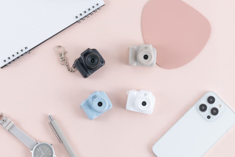 Cuatro pequeñas cámaras de juguete en negro, gris, azul y blanco están dispuestas sobre una superficie rosa, junto a un bloc de notas, un reloj gris, un bolígrafo, una alfombrilla para ratón y un teléfono inteligente blanco.