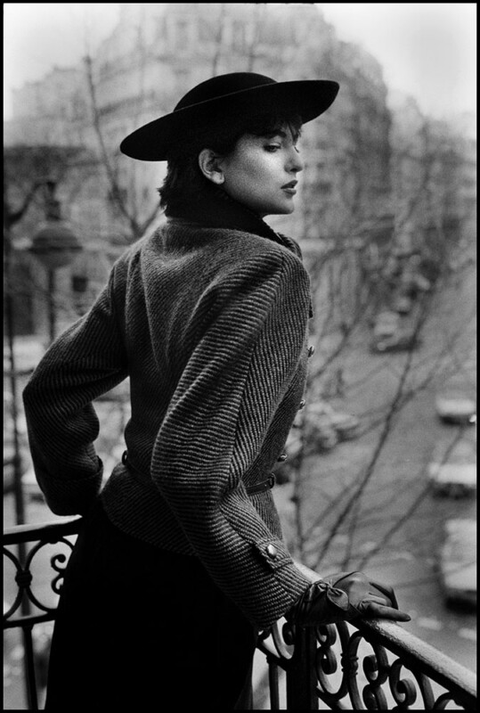 A woman in a vintage outfit and wide-brimmed hat stands on a balcony, looking over her shoulder with eyes closed. The background shows a blurred city street with cars and bare trees. The image is in black and white.