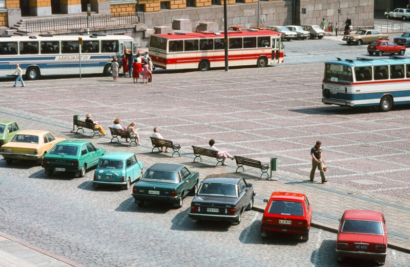 Una plaza abierta con coches aparcados y algunos bancos, algunos de los cuales estaban ocupados por gente. Al fondo hay tres autobuses aparcados y alrededor de la plaza hay varias personas paseando o de pie. El suelo está pavimentado con piedras estampadas.