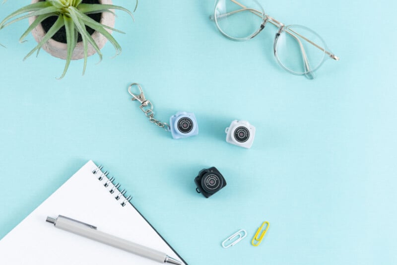 Cuaderno de espiral plano con bolígrafo, dos llaveros de mini cámara, gafas, clips y una pequeña planta en maceta sobre fondo azul claro.