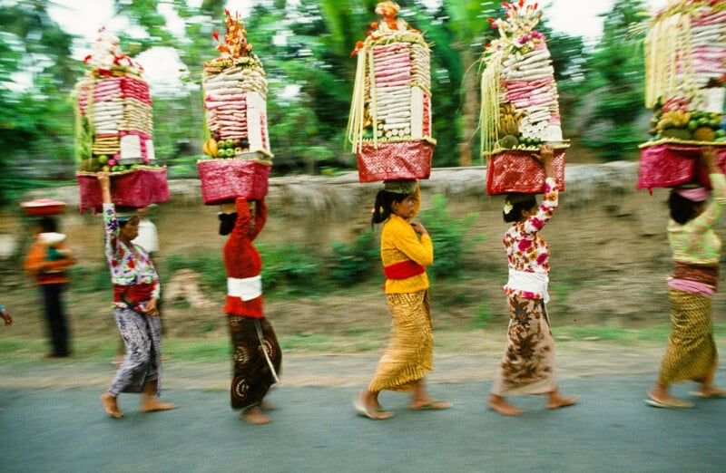 Un grupo de mujeres descalzas, vestidas con trajes tradicionales de colores brillantes y cada una con una ornamentada cesta de sacrificio en la cabeza, caminaban en fila por un camino rural bordeado de árboles.