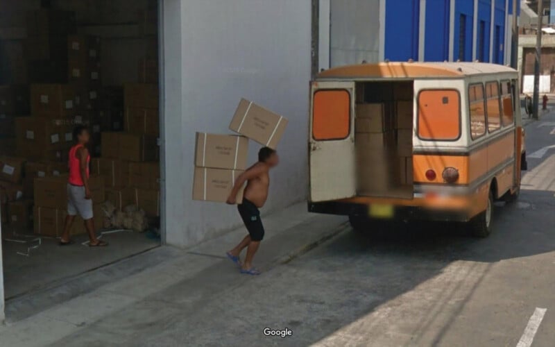 Un hombre sin camisa llevaba una pila de cajas desde el almacén hasta una furgoneta naranja estacionada afuera. Otro hombre se encontraba en la puerta del almacén, rodeado de más cajas.