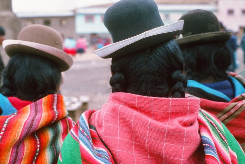 Vista desde atrás de tres mujeres vestidas con coloridos mantones andinos tradicionales y bombines. Su cabello oscuro estaba trenzado y parecían estar en un pueblo al aire libre, con edificios borrosos en el fondo.