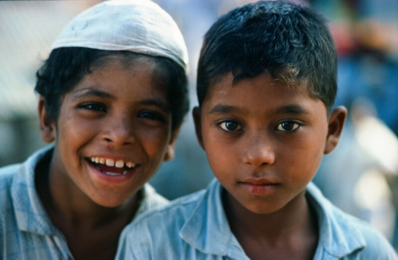 Dos niños pequeños estaban uno cerca del otro, uno con una amplia sonrisa y el otro con una expresión neutra. Ambos vestían camisas de colores claros y uno de los niños llevaba un sombrero blanco. El fondo es borroso y al aire libre.