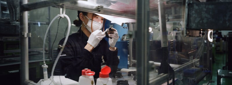 A person wearing a face mask and gloves carefully inspects an object under bright lights in a factory or laboratory setting surrounded by various equipment and containers.