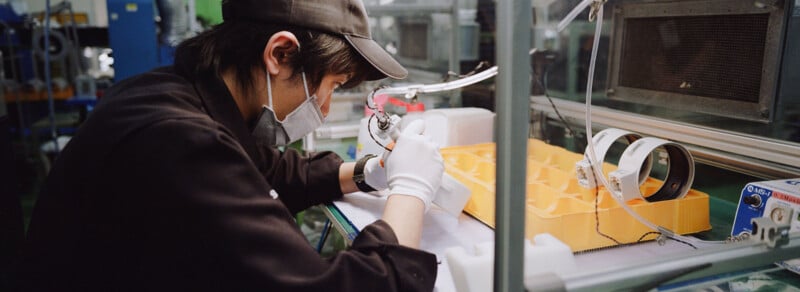 A person wearing a mask, gloves, and a cap works with small tools at a workstation, assembling or inspecting items next to a yellow tray and electronic equipment in a factory setting.