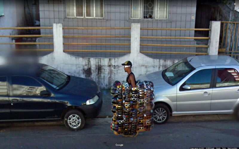 Un vendedor ambulante camina entre coches aparcados cubiertos con gafas de sol, vendiéndolos en el casco urbano, con un edificio de azulejos al fondo.