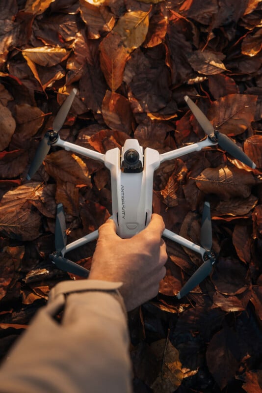 La mano de un hombre sostiene un dron blanco con cuatro hélices en el suelo cubierto de hojas marrones de otoño.