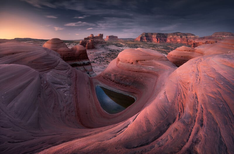 Al atardecer, el sorprendente paisaje desértico presenta formaciones arremolinadas de arenisca roja y una pequeña piscina en forma de corazón ubicada entre las rocas, con mesas distantes y un cielo brillante al fondo.