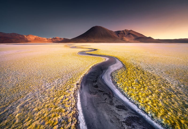Un arroyo oscuro y serpenteante atraviesa una vasta extensión de salinas amarillas y blancas, que conducen a montañas distantes bajo un espectacular cielo de atardecer en profundos tonos azules y naranjas.