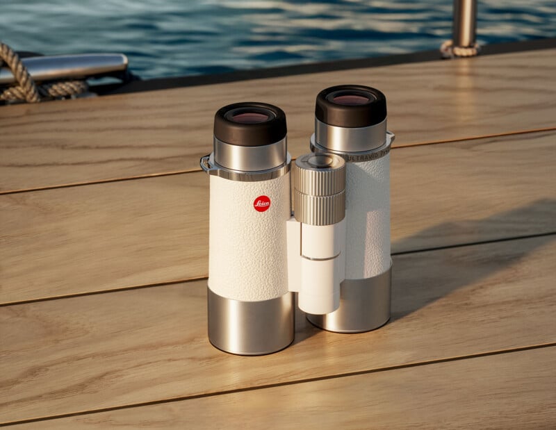A pair of white Leica binoculars rests on a wooden surface near a body of water, with sunlight reflecting off the water and the deck.