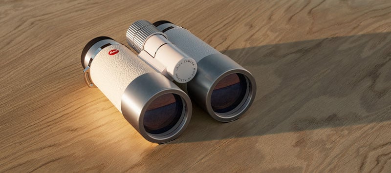 A pair of white binoculars with black eyepieces and a silver focus wheel rests on a wooden surface, casting a soft shadow in warm lighting.