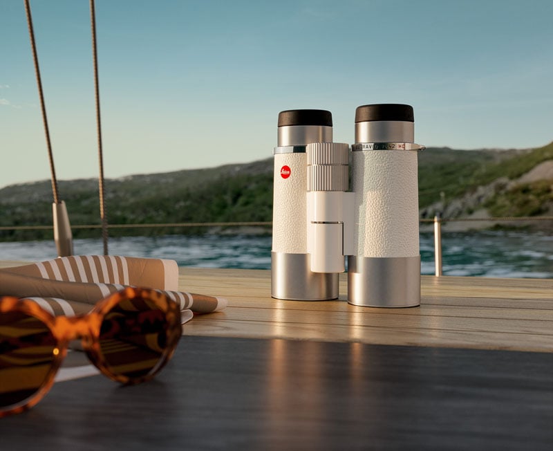 A pair of white binoculars sits on a wooden surface of a boat, with water and a hilly shoreline in the background. Sunglasses and a striped cloth are nearby, evoking a relaxing outdoor scene.