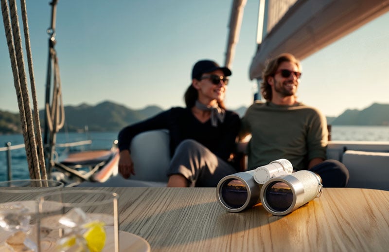 A pair of binoculars sits on a table in the foreground, while a smiling couple relaxes together on a sailboat with mountains and water visible in the background.