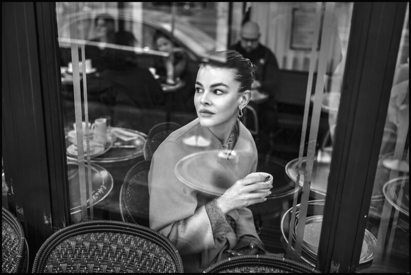 A woman sits at a café table by a window, holding a small cup and looking thoughtfully to the side. Several empty cups and plates are on the tables around her, with other people visible in the background. The photo is in black and white.