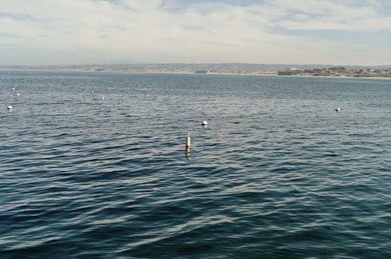 Calm ocean water with gentle waves, several floating buoys, and a coastline with hills and buildings in the distance under a partly cloudy sky.