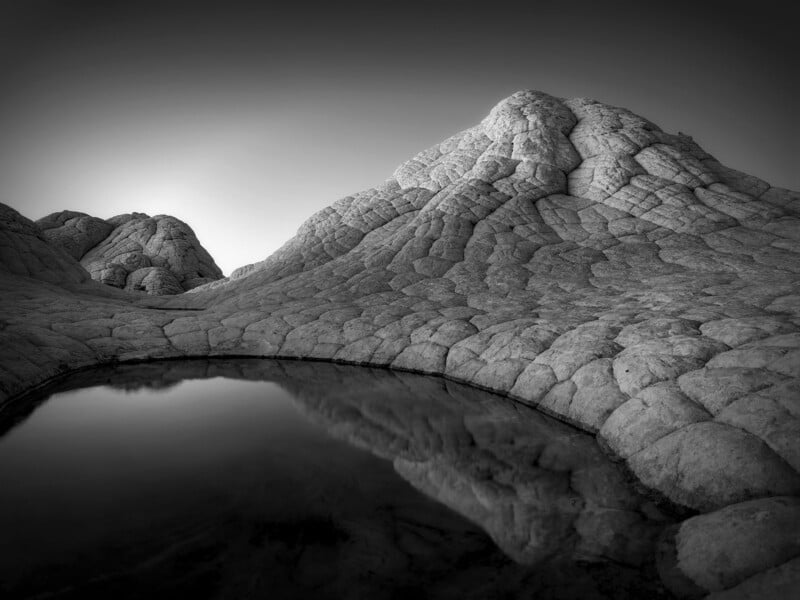 La fotografía en blanco y negro muestra una montaña rocosa y texturizada con un pequeño estanque tranquilo en primer plano, que refleja la superficie de la montaña. Bajo un cielo despejado, el paisaje parece sobrenatural y árido.