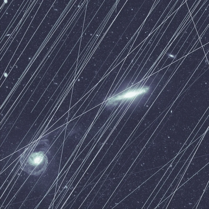 A starry night sky with two bright galaxies, streaked by numerous diagonal white lines from satellite trails crossing the image.