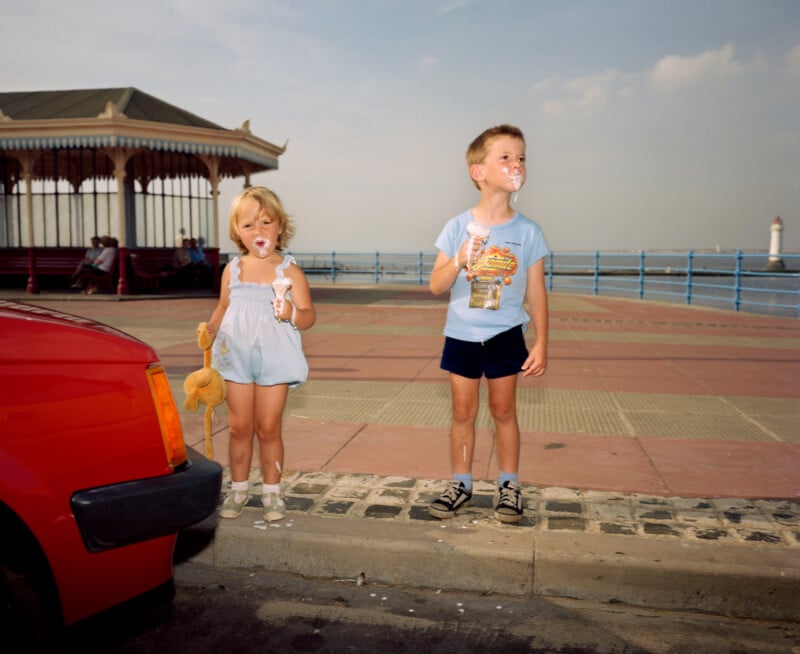 Two young children stand on a promenade by the sea, eating ice cream with messy faces. A red car is parked nearby, and a lighthouse and pavilion are visible in the background. The girl holds a teddy bear.
