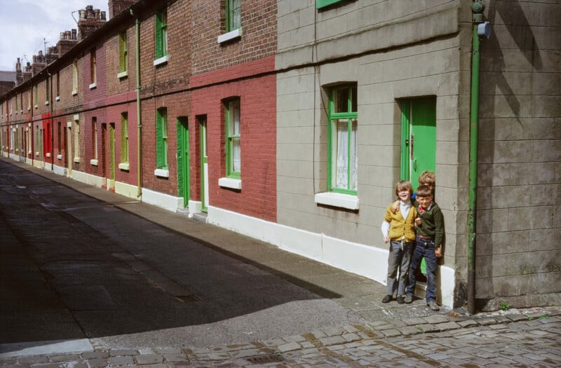 Los tres niños estaban juntos en una tranquila calle adoquinada, frente a una hilera de casas de ladrillo con puertas y ventanas pintadas de verde brillante. Esta escena parece ser de los últimos diez años.