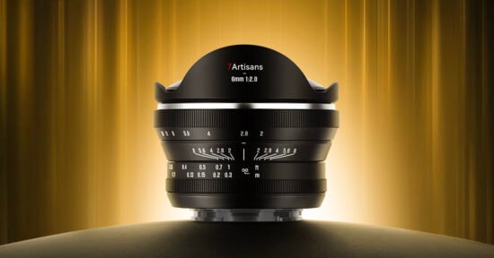 A 7Artisans 6mm f/2.0 camera lens is displayed upright on a dark surface, with a glowing golden background and vertically streaked light, highlighting its wide, curved front element and detailed focus markings.