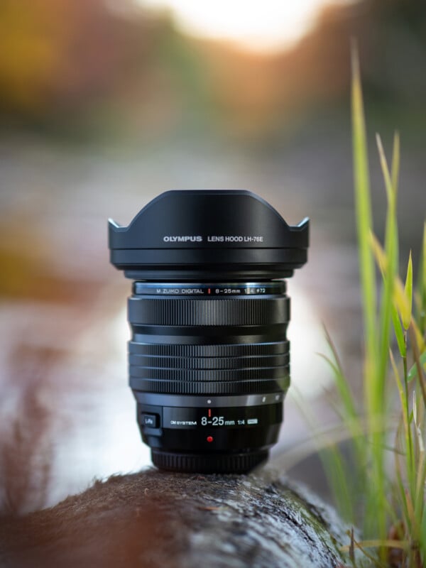 A black Olympus camera lens with a lens hood stands upright on a wet rock outdoors, surrounded by blurred natural greenery and soft, colorful light in the background.