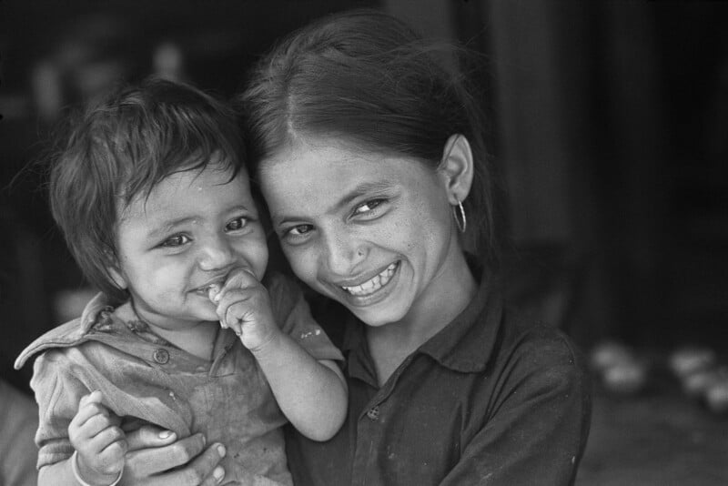 Una niña sonriente sostiene a un niño en sus brazos. Ambos miran a la cámara; el niño se chupa el dedo, mientras la niña lo abraza y lleva aretes. La imagen está en blanco y negro.