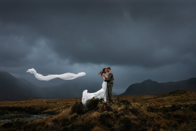 La novia y el novio se abrazan en una colina cubierta de hierba bajo un espectacular cielo oscuro. El velo de la novia ondea con el viento, quedando muy atrás, con las montañas al fondo.