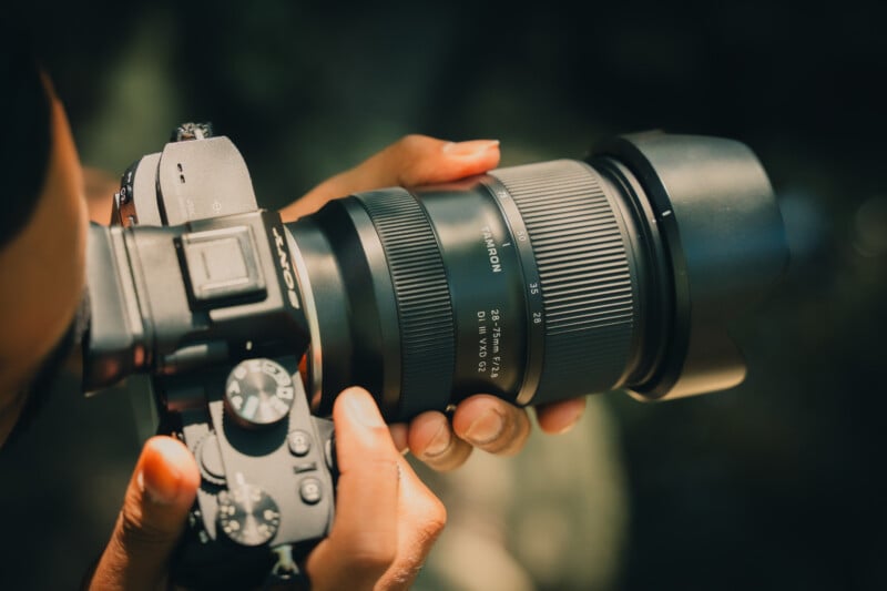 Un hombre sostiene una cámara Sony con un gran teleobjetivo Tamron, ajustando la configuración. La cámara y la lente están enfocadas y el fondo exterior está borroso.