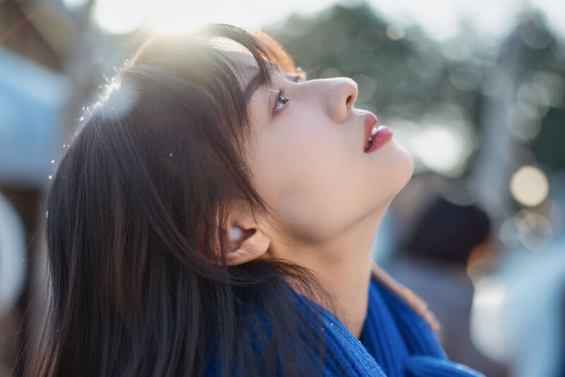 A young woman with long dark hair looks upwards, illuminated by sunlight behind her. She wears a blue scarf and appears thoughtful, with a soft background of trees and bokeh light.