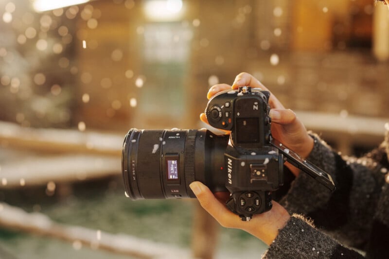 Close-up of a person holding a Nikon Z series camera with a large lens, outdoors in soft sunlight with falling snowflakes and a blurred background.