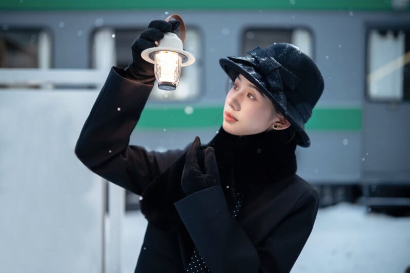 A woman wearing a black coat, gloves, and hat with a bow holds a glowing lantern while standing outside in light snowfall near a train carriage.