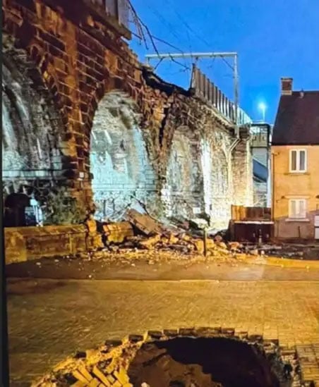 Un viaducto de ladrillo dañado con una pared parcialmente derrumbada por la noche, escombros esparcidos por el suelo y un gran agujero en primer plano cerca de las casas cercanas.