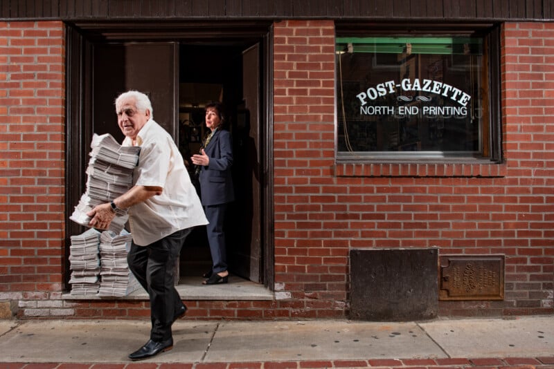 An older man carries a large stack of newspapers outside a brick building labeled "Post-Gazette North End Printing" as a woman stands in the doorway behind him.