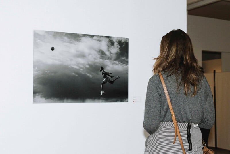 A person with long hair, seen from behind, observes a black-and-white photograph of a person jumping to kick a soccer ball against a cloudy sky, displayed on a white gallery wall.