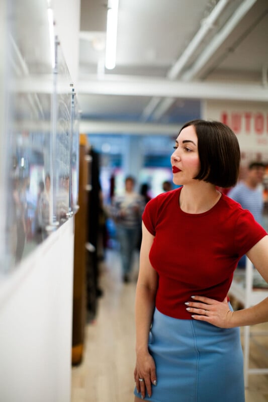 Una mujer con cabello corto y oscuro, vestida con una blusa roja y una falda azul claro, está parada en el interior y mira atentamente la obra de arte en la pared, con gente y luces brillantes al fondo.